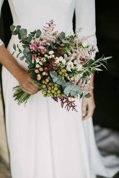 a woman in a white dress holding a bouquet of flowers and greenery on her wedding day