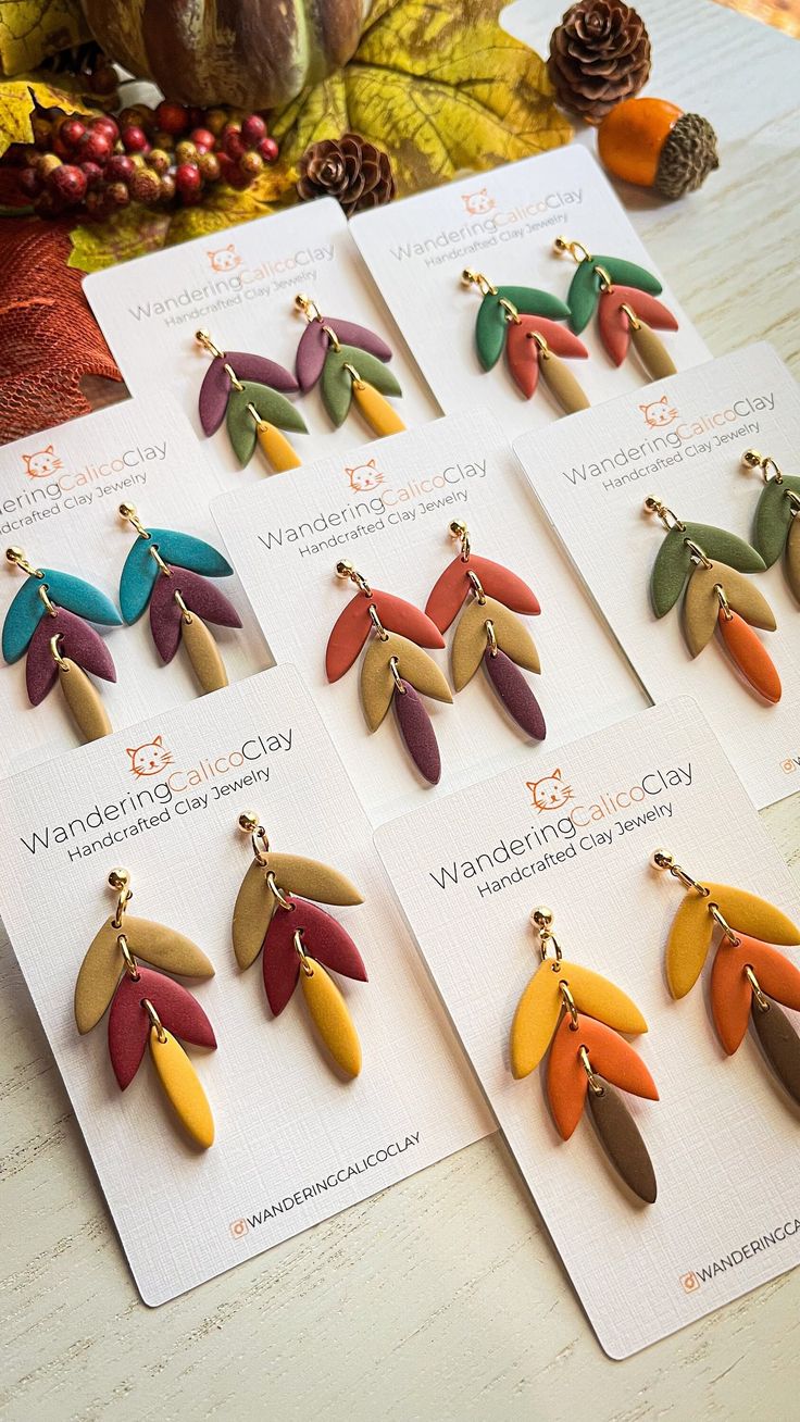 six pairs of earrings with colorful leaves on them sitting on a table next to some pine cones