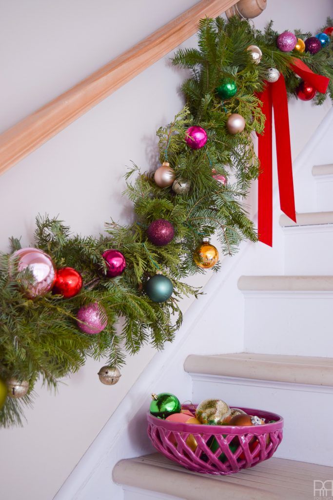 a christmas garland on the stairs with ornaments in a pink basket and red ribbon hanging from the banister