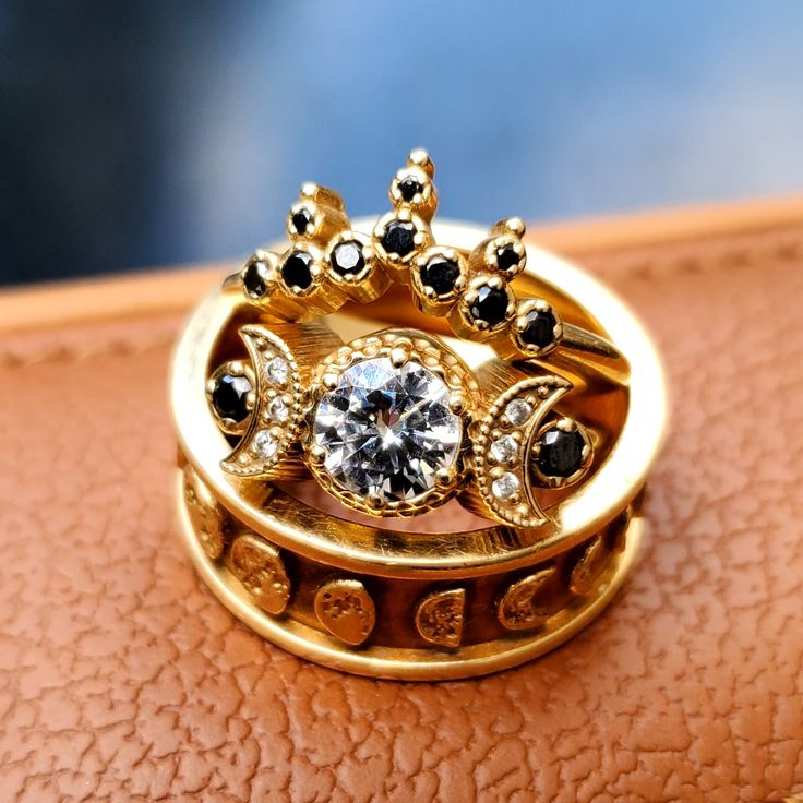 a gold ring with black and white stones on it sitting on top of a brown leather surface