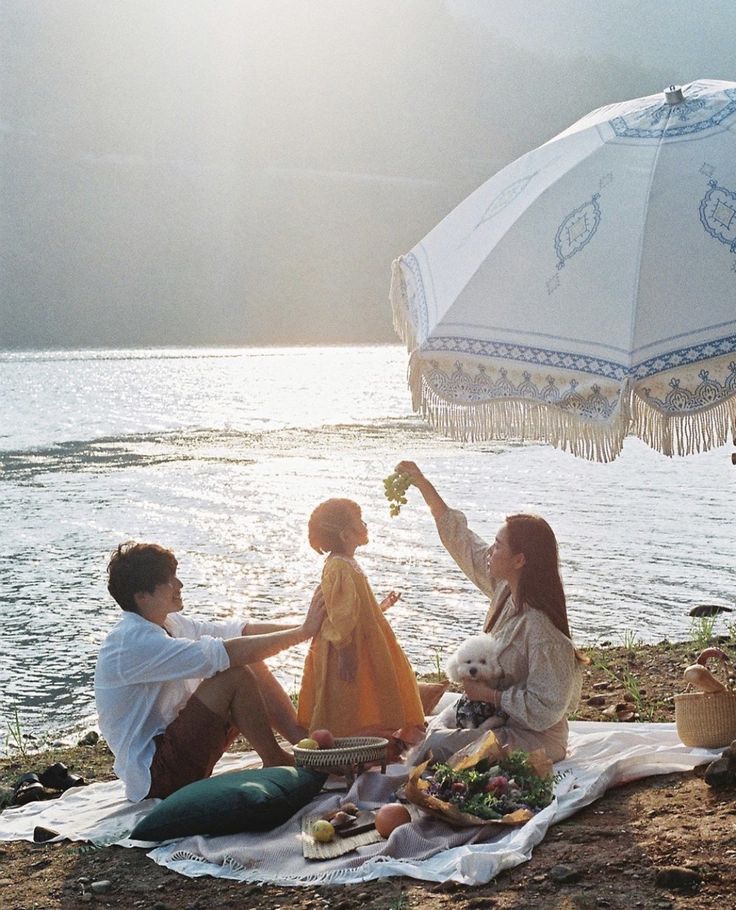 two women and a man sitting on the beach with an umbrella over them, eating food