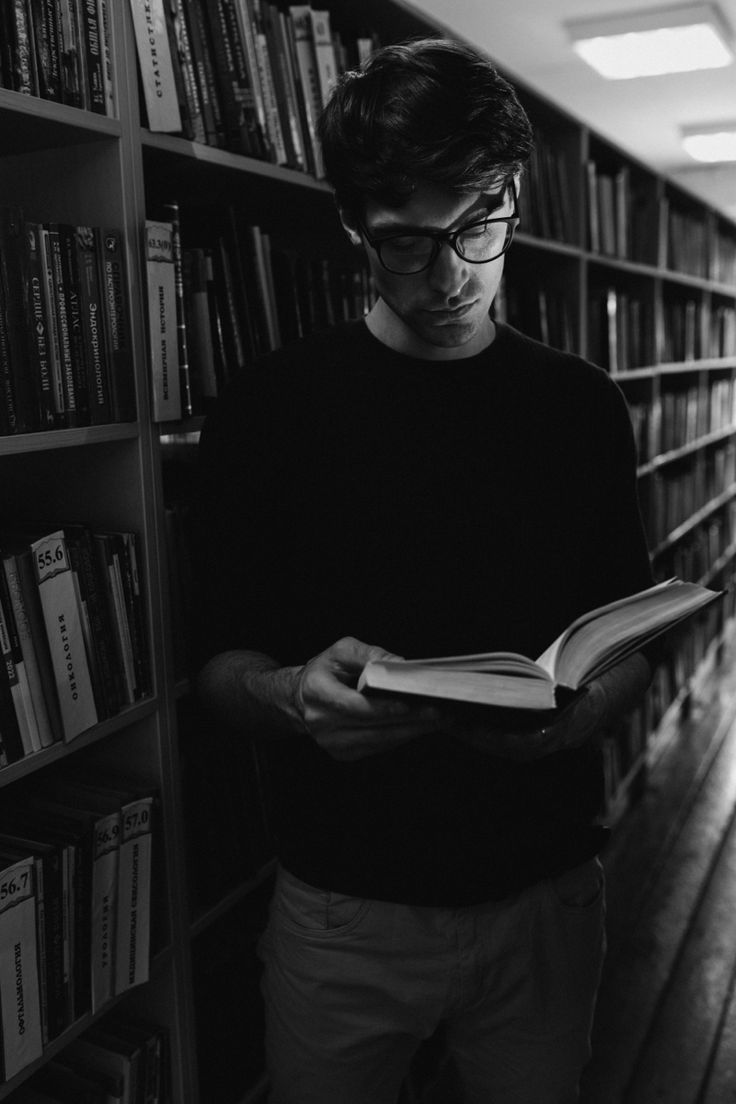 a man standing in front of a bookshelf holding an open book and looking at it