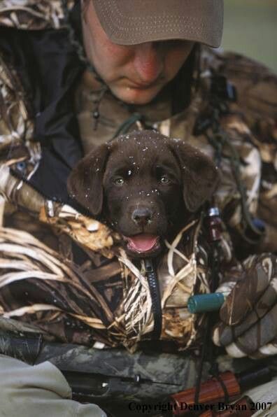 a man holding a dog in his arms while wearing a hat and hunting gear on the ground