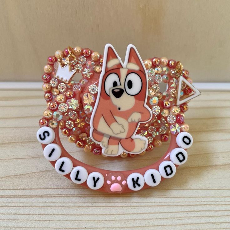 a pink and white dog brooch sitting on top of a wooden table next to a name tag