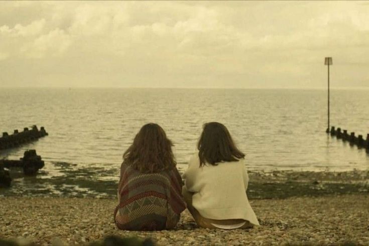 two women sitting on the beach looking out at the water