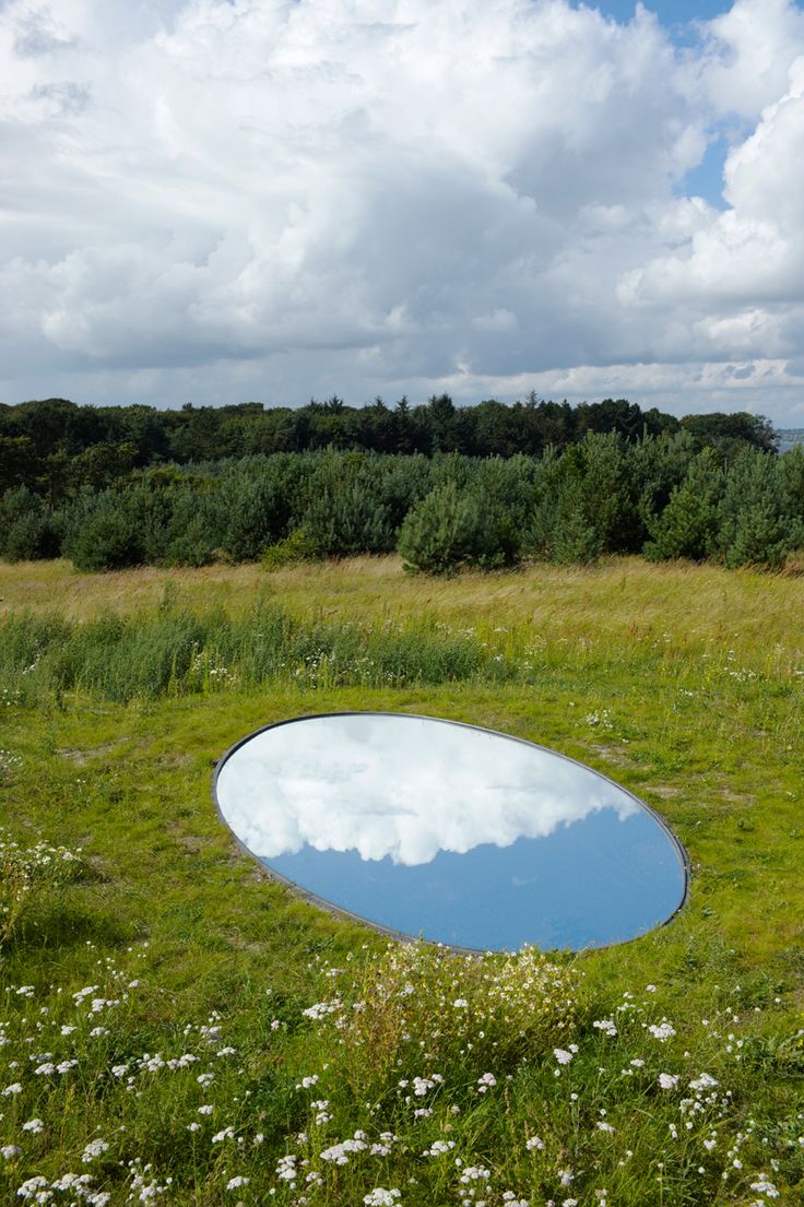 a large mirror in the middle of a grassy field with trees and clouds reflected in it