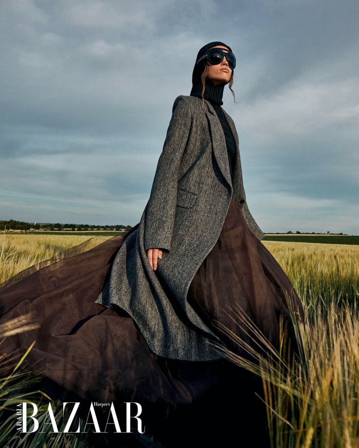 a woman is standing in the middle of a field wearing a gray coat and black dress