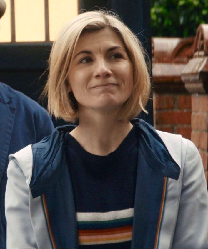 a woman with blonde hair wearing a blue jacket and striped shirt is standing in front of a brick building