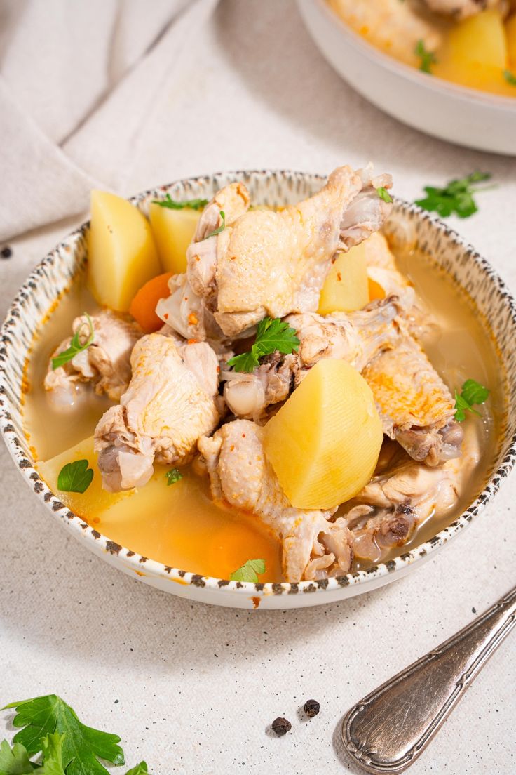 a bowl filled with chicken and potatoes on top of a white table next to a spoon