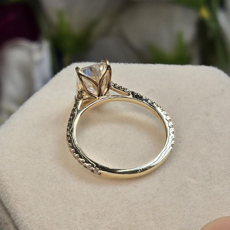 a diamond ring sitting on top of a white box with flowers in the background and someone's hand holding it