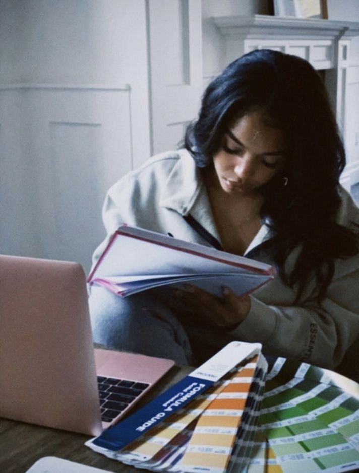 a woman sitting in front of a laptop computer holding a book and looking at it