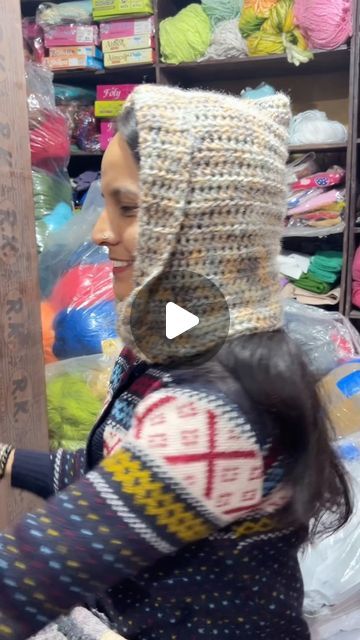 a woman wearing a crocheted hat in a store with balls of yarn behind her