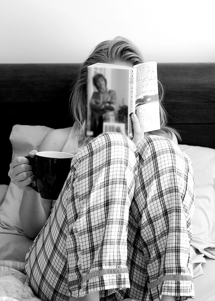 a woman laying in bed reading a book and holding a cup with her feet up