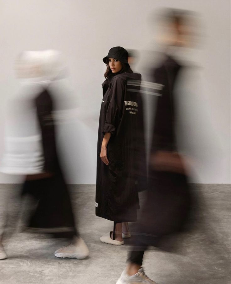 a woman standing in front of a white wall with blurry people walking around her