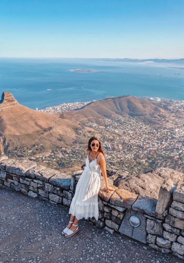 a woman standing on top of a stone wall next to the ocean with mountains in the background