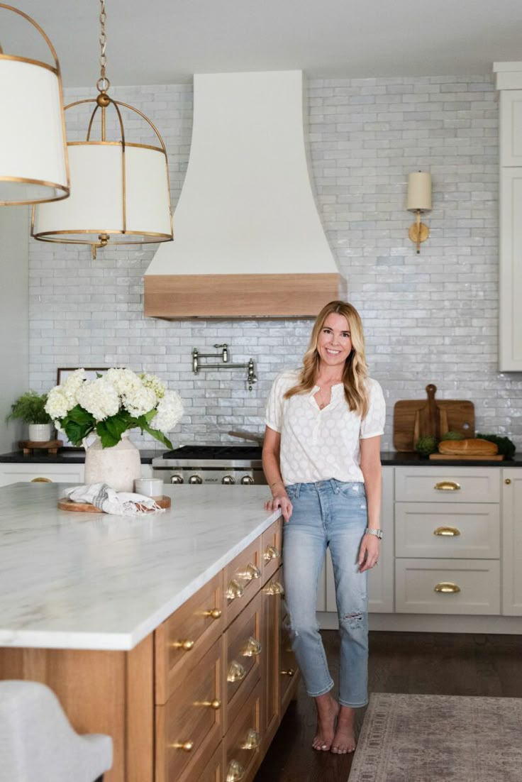 a woman standing in the middle of a kitchen with white cabinets and marble counter tops