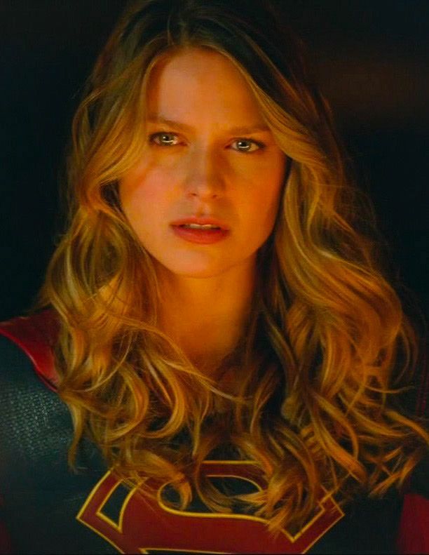 a woman with long blonde hair wearing a superman costume and looking at the camera while standing in front of a dark background