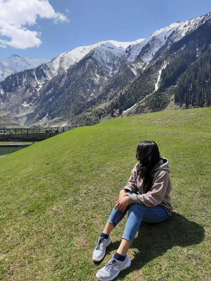 a woman sitting on top of a lush green field next to mountains covered in snow