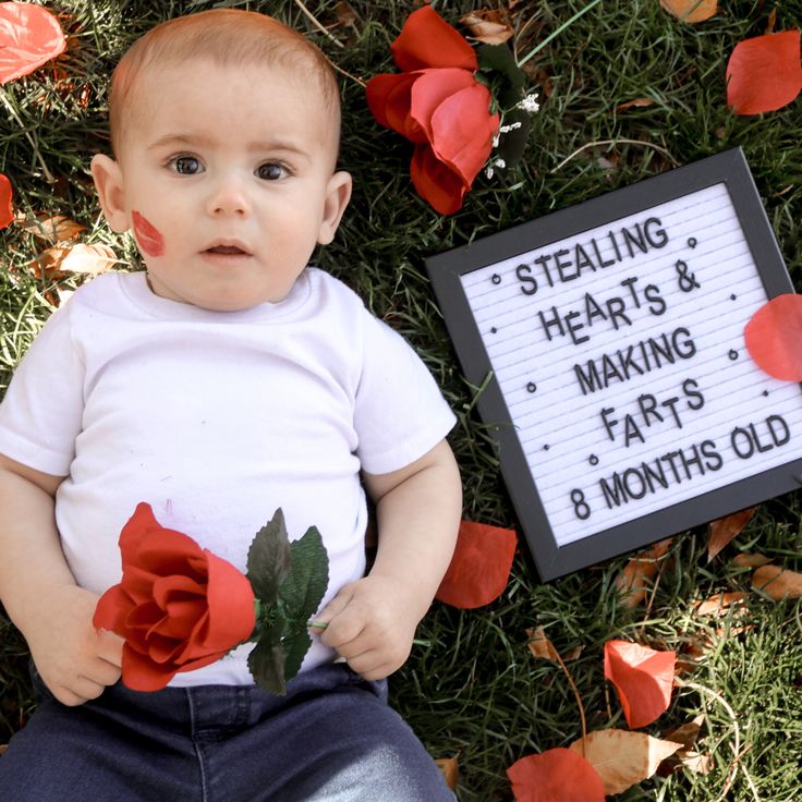 a baby laying in the grass next to a sign that says stealing hearts and making parts 8 months old