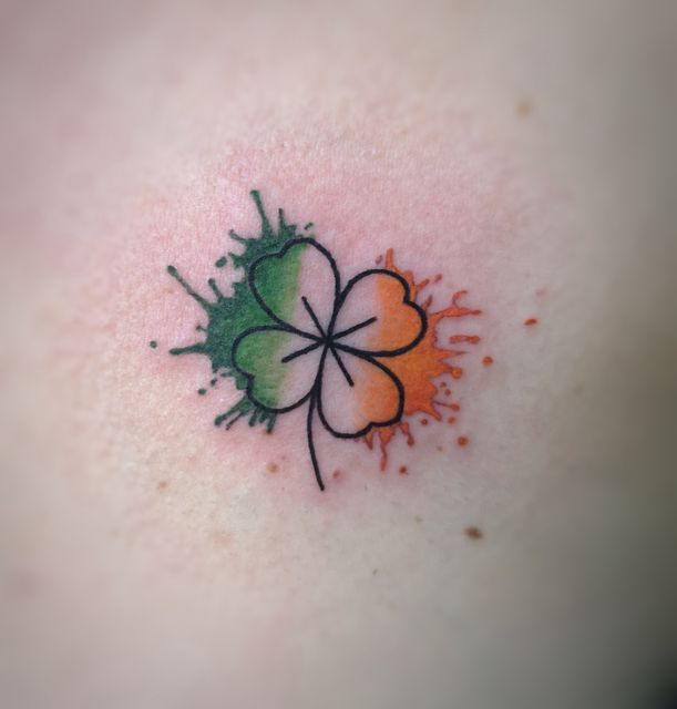 a four leaf clover tattoo on the back of a woman's chest, with orange and green ink splatters all over it