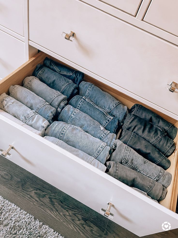 an open drawer filled with jeans on top of a carpeted floor next to a white dresser