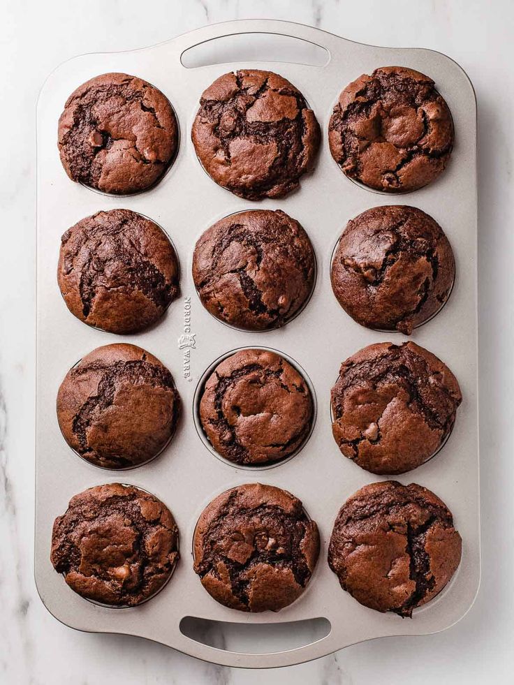 twelve chocolate muffins in a baking pan on a marble counter top, ready to be eaten