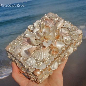 a hand holding a box with seashells and pearls on the beach next to the ocean