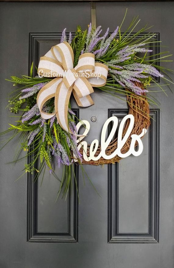 a wreath with the word hello written on it hanging on a gray front door, decorated with lavenders and greenery