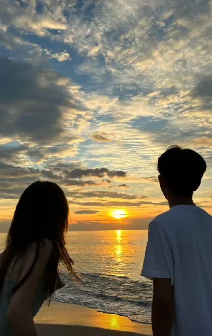 two people are standing on the beach watching the sun go down over the water and clouds