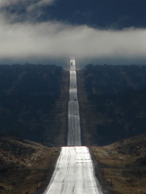 an empty road with no cars on it in the middle of mountains and clouds above