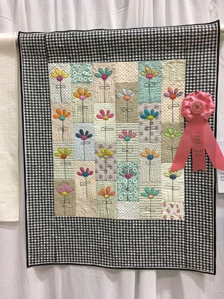 a pink teddy bear sitting on top of a quilted wall hanging in front of a white curtain