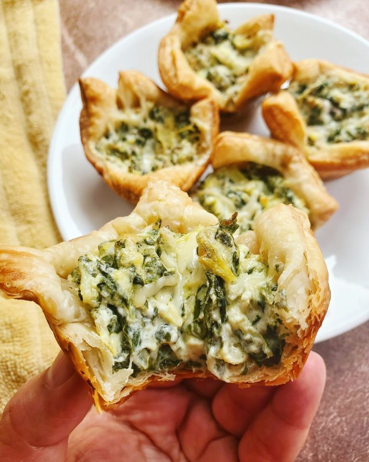 a hand holding a small pastry with spinach and cheese in it on a plate