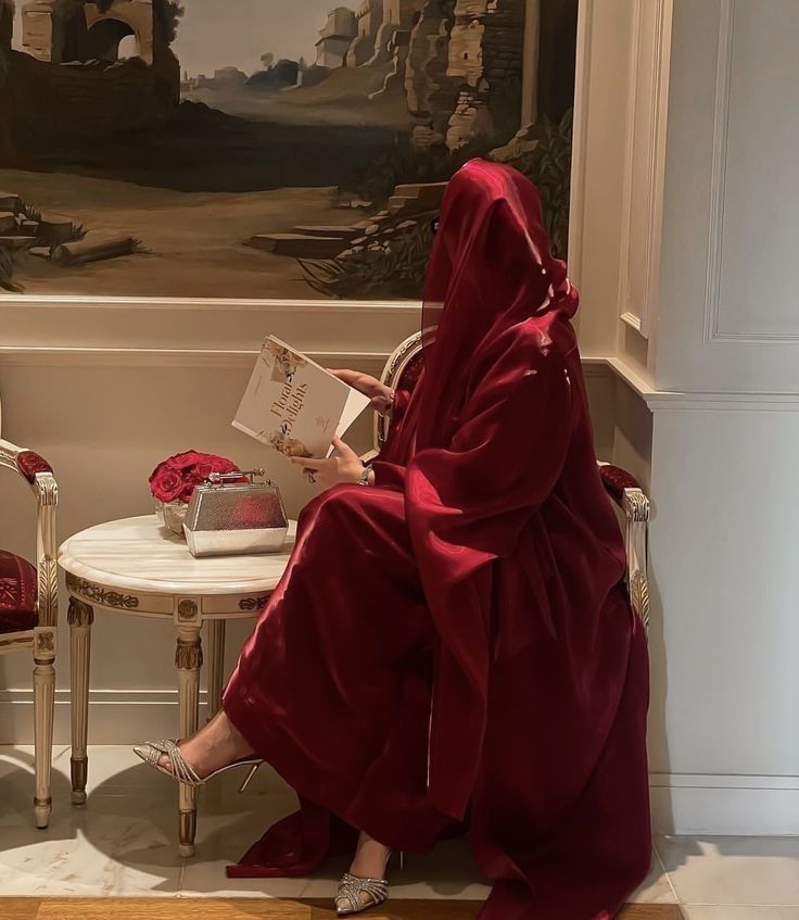 a woman in a red robe is reading a book near a table with two chairs and a painting on the wall