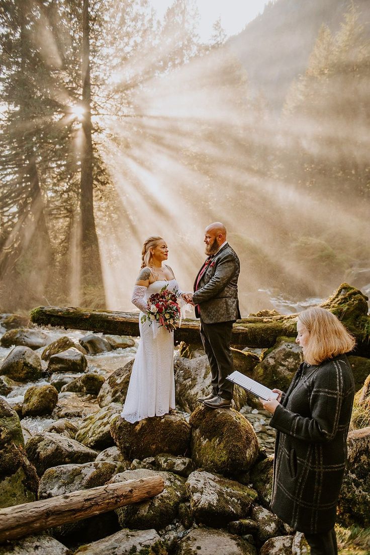 a bride and groom are standing on rocks in the woods with sunlight coming through the trees