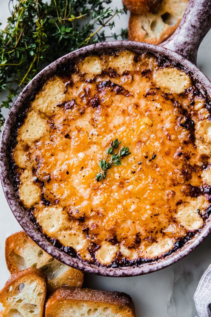 a casserole dish with cheese and bread on the side, surrounded by fresh herbs