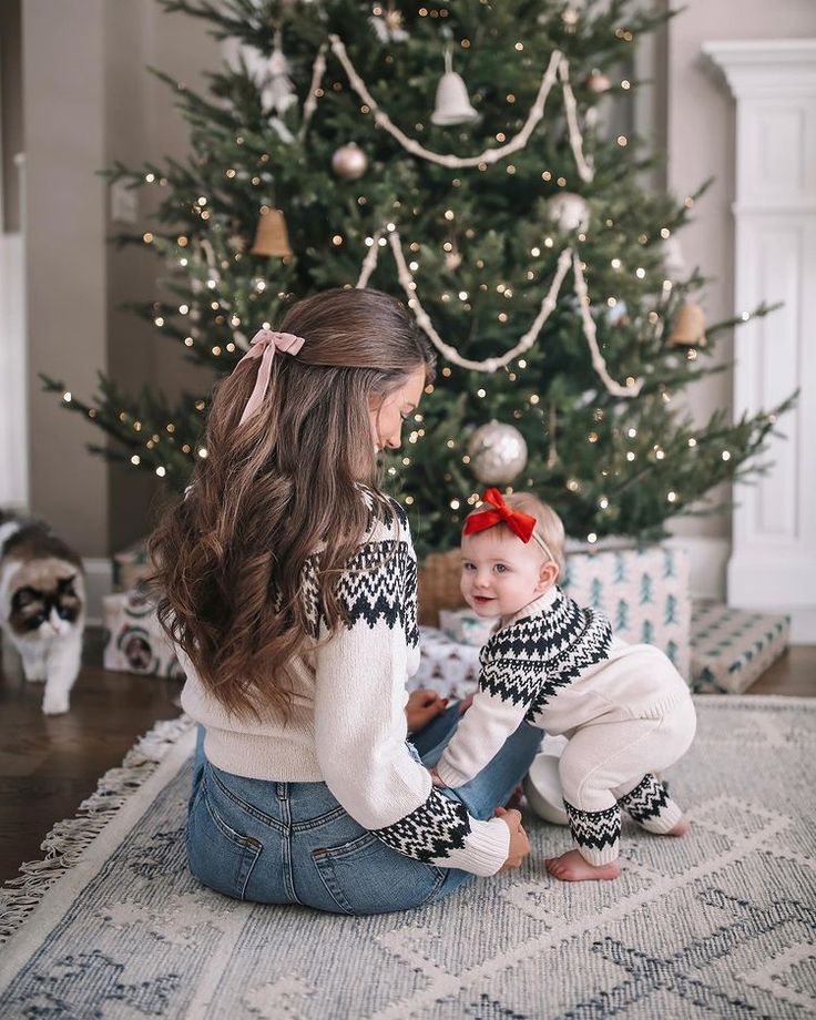 a woman sitting on the floor next to a christmas tree with her baby in front of it