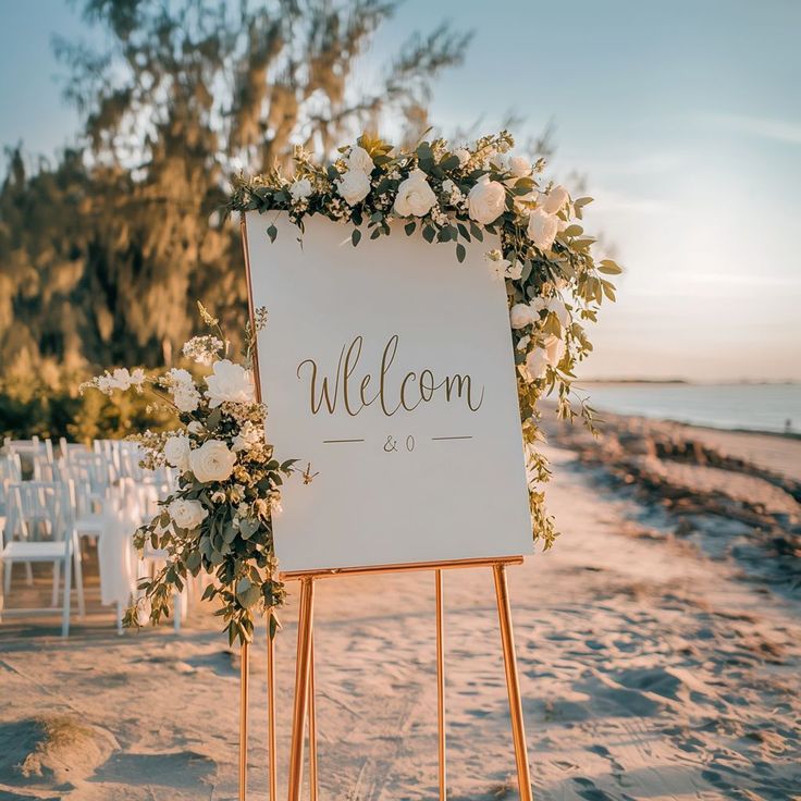 a welcome sign on the beach with white flowers and greenery is displayed for an outdoor wedding ceremony