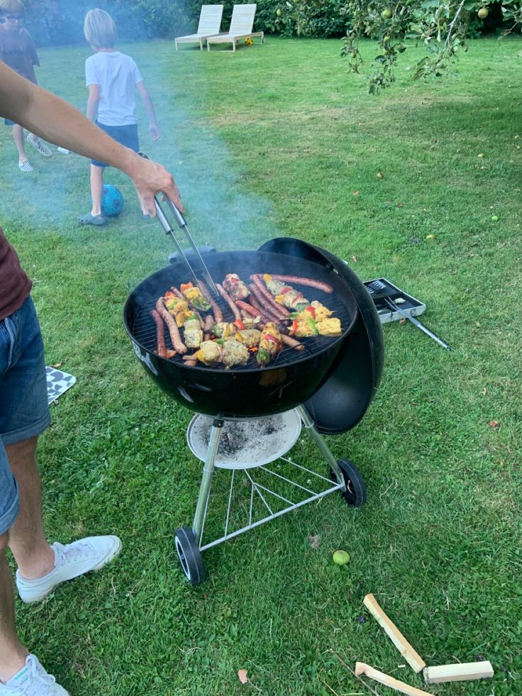 a man grilling food on top of a bbq in the grass next to other people