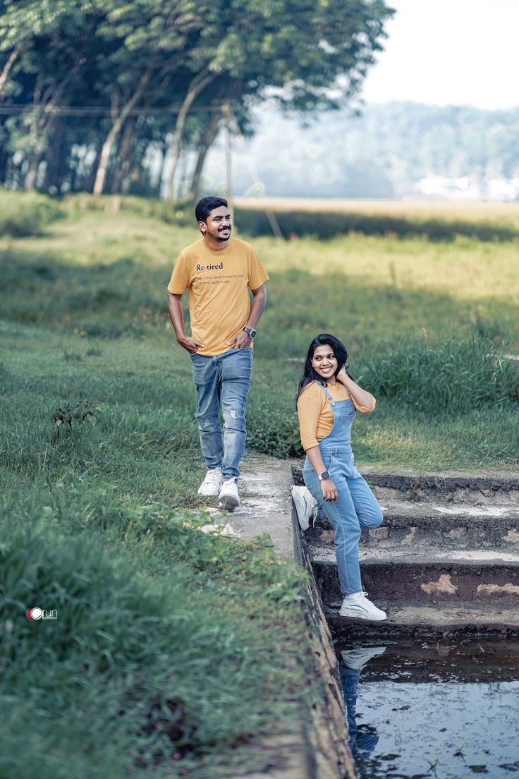 two people standing next to each other on the side of a river with grass and trees in the background