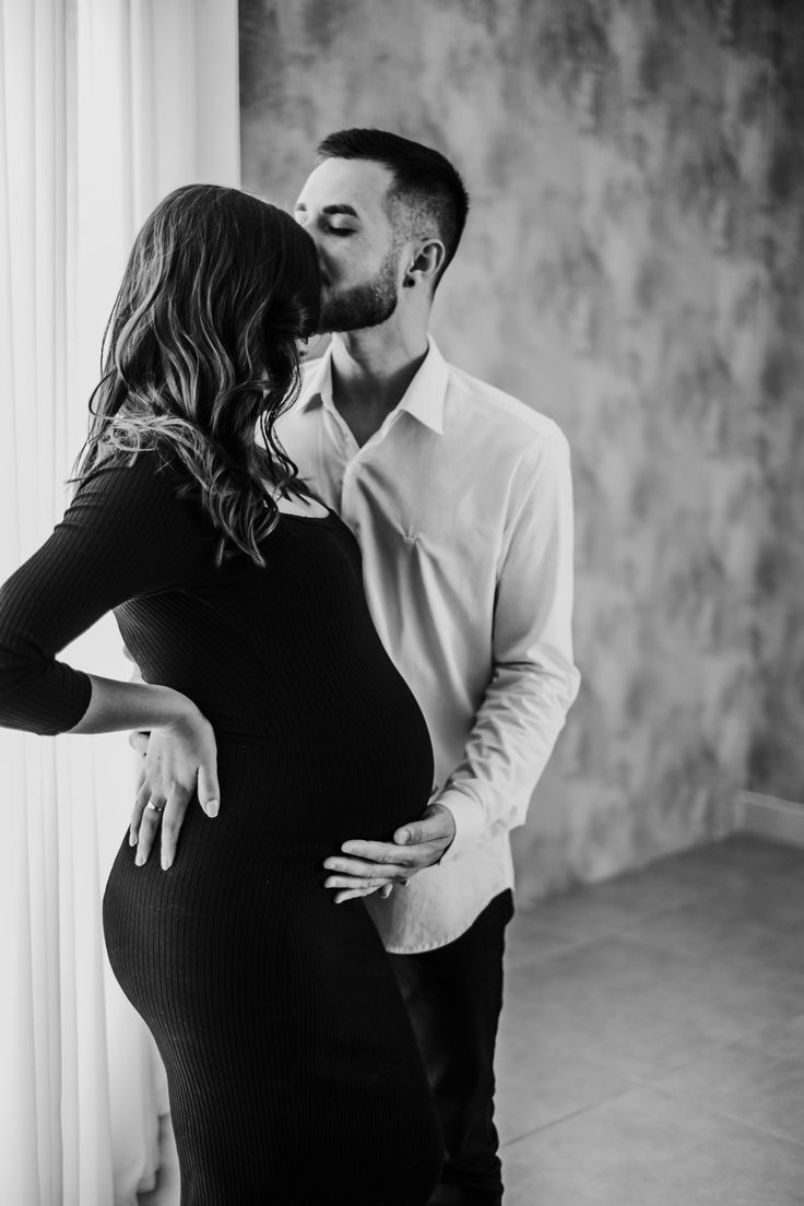 black and white photo of a pregnant woman kissing her husband's forehead in front of a window