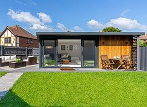 a modern house with an open patio and wooden furniture on the lawn in front of it