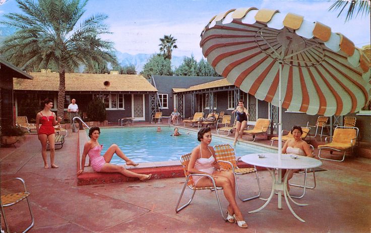 several women sitting around a swimming pool with an umbrella over the pool and chairs in front of it