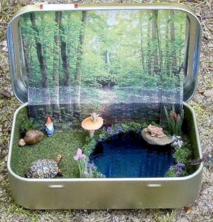 an open tin box with miniature people in the woods surrounding it on grass and rocks