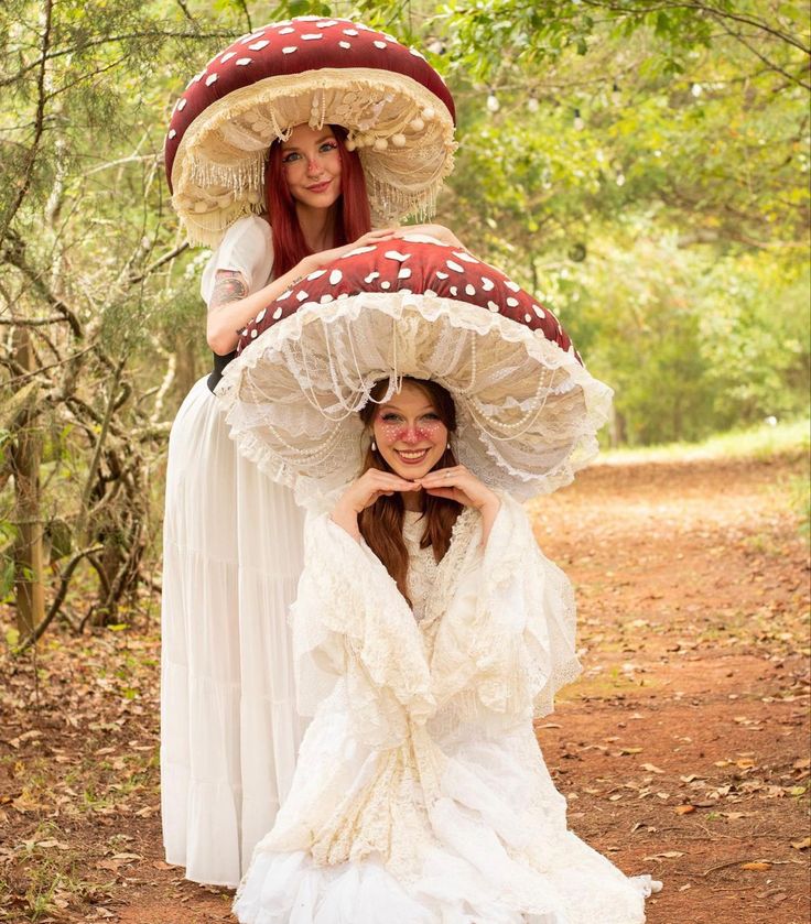 two women dressed in white dresses and large mushroom hats pose for the camera while standing on a dirt path