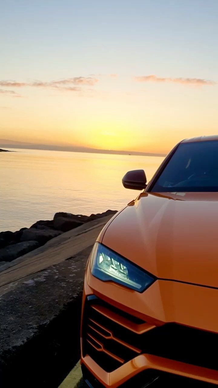 an orange sports car parked on the side of the road next to the ocean at sunset