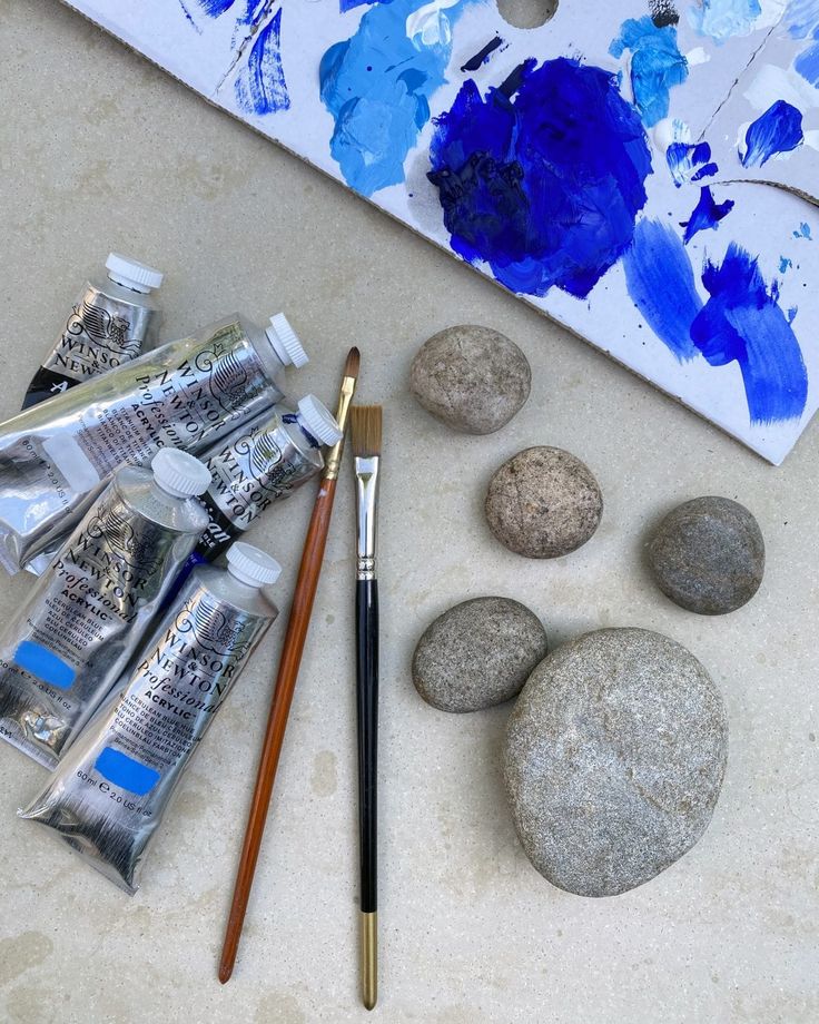 some rocks and paint are sitting on the ground next to a brush, pencil, and watercolors