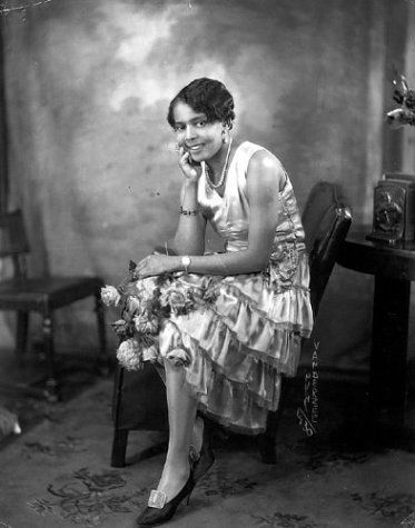 an old photo of a woman sitting on a chair with flowers in her hand and smiling at the camera