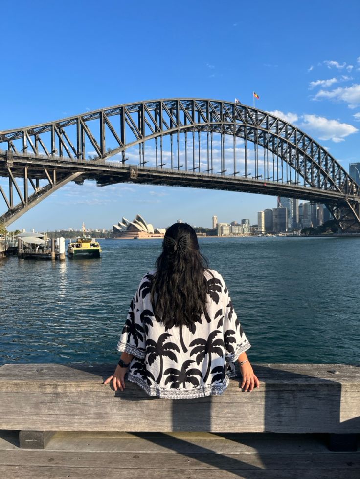 a woman sitting on a bench looking out at the water and a bridge in the background