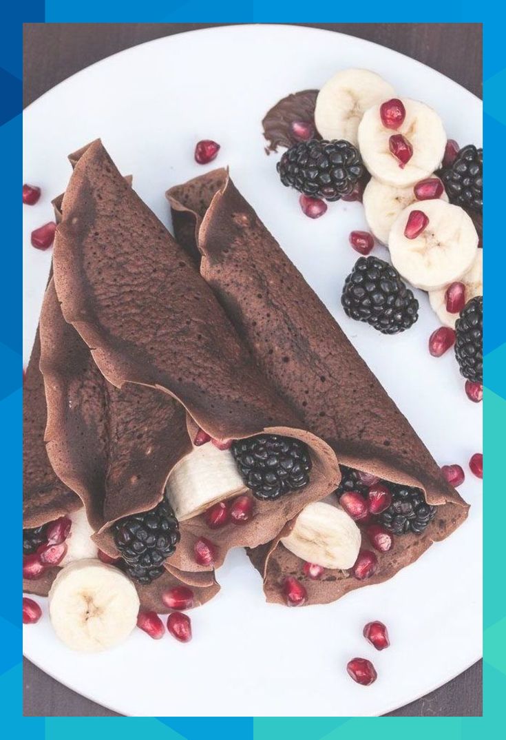 a white plate topped with chocolate crepes covered in bananas and pomegranates