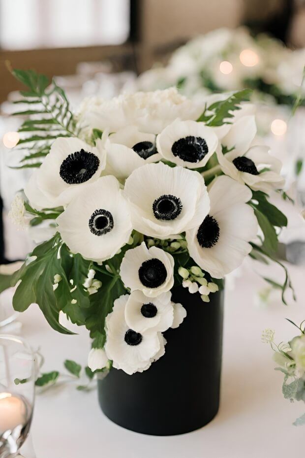 an arrangement of white flowers in a black vase on a table with candles and greenery